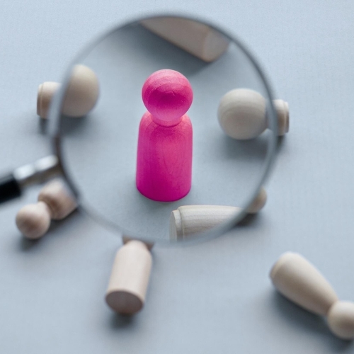 Magnifying glass highlighting a single pink game piece among neutral pieces, symbolizing pinpointing the right hire.